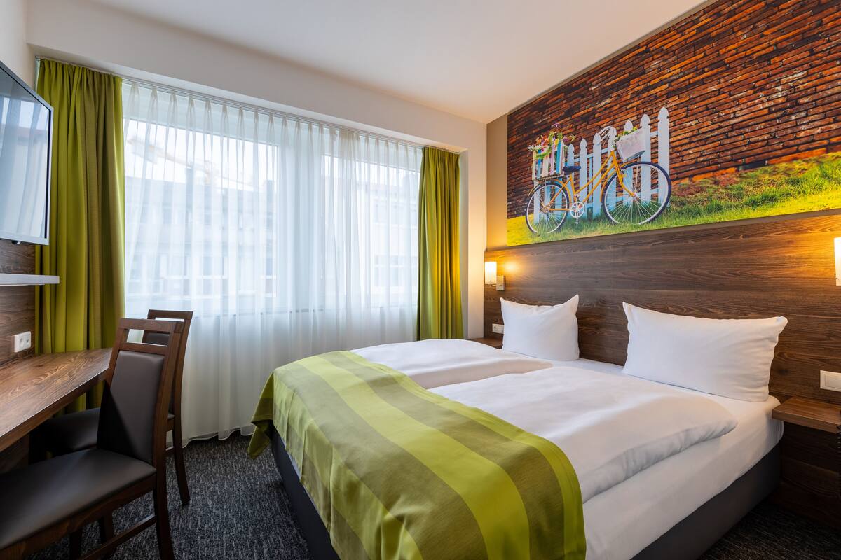 A hotel room with a bed, a desk, and a chair. There is a window with curtains, a painting on the wall, and a wooden headboard.