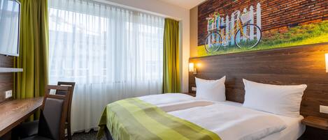 Habitación de hotel con cama, escritorio y silla. Se observa una ventana con cortinas, un cuadro en la pared y un cabecero de madera.