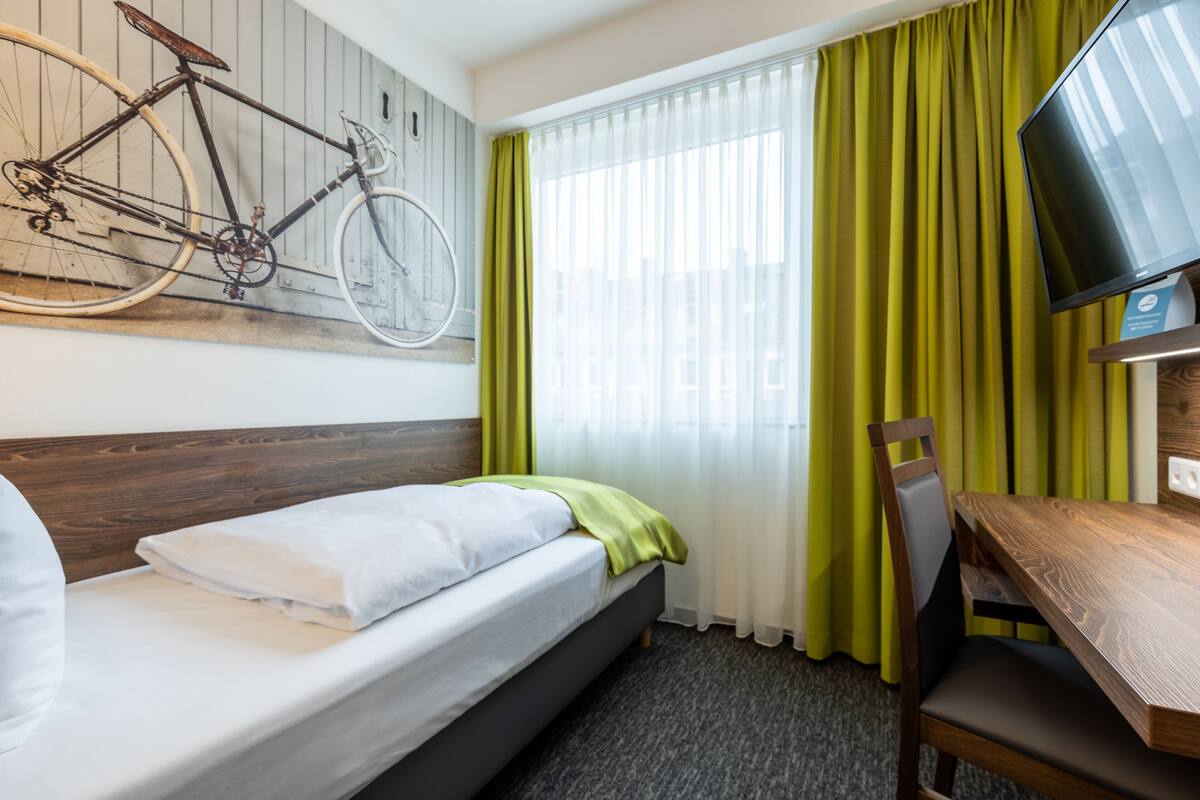 A hotel room with a bed, a wooden desk, a television, a bicycle, and a window with curtains.