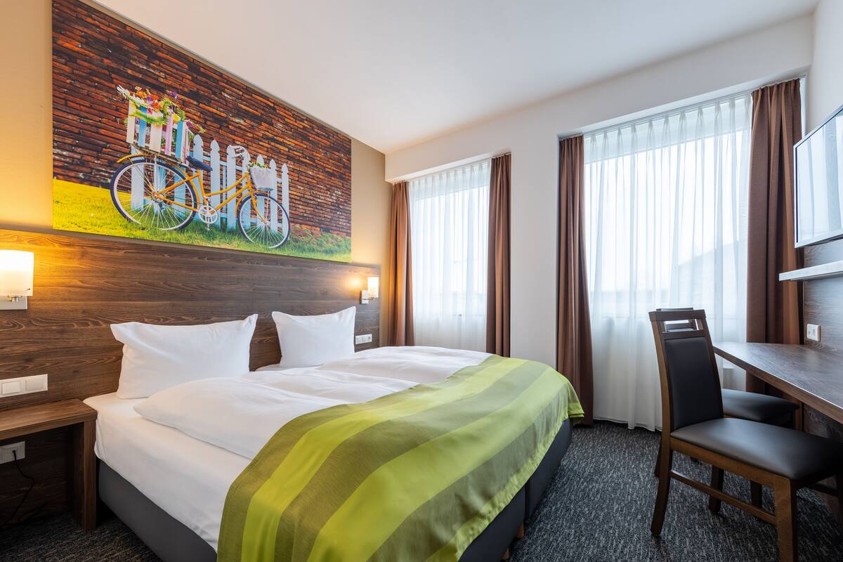 A hotel room with a large bed, a desk, a chair, and a painting of a bicycle on the wall.