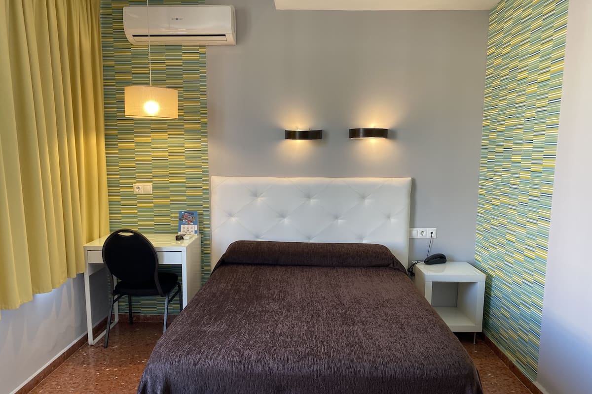 A hotel room with a bed, a desk, a chair, an air conditioner, and two wall-mounted lights.