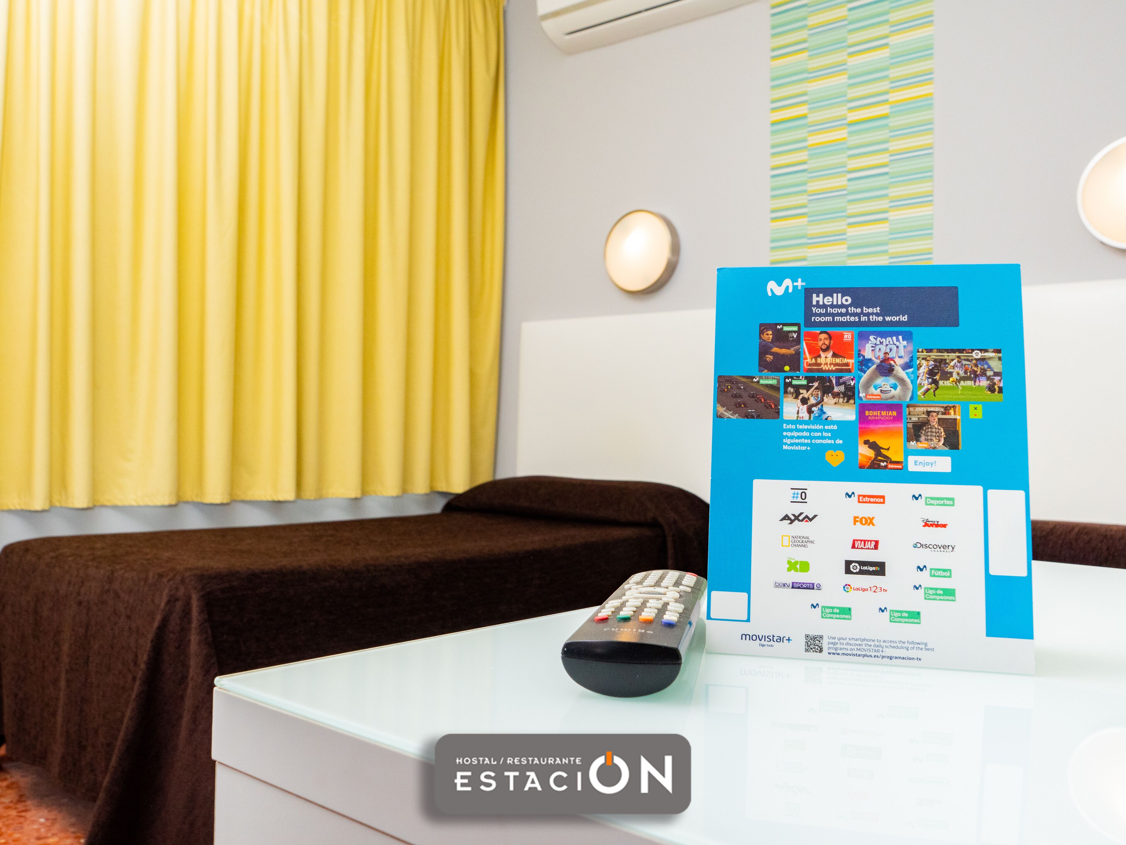 A hotel room with a bed, a remote control, and a Movistar+ TV stand displaying various channels and logos.