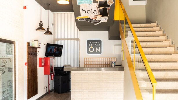 Un hall d'hôtel comprenant un comptoir d'accueil, un escalier et une enseigne fixée au mur sur laquelle on peut lire : « Hostal Estaci ON ».