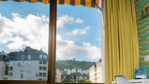 Une pièce dotée d’une grande fenêtre donnant sur un paysage urbain, avec un rideau jaune et une vue sur des immeubles et une colline.
