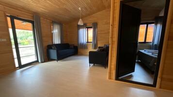 Una habitación amplia con paredes y suelos de madera, un sofá negro, un sillón pequeño y una cama con colcha blanca.