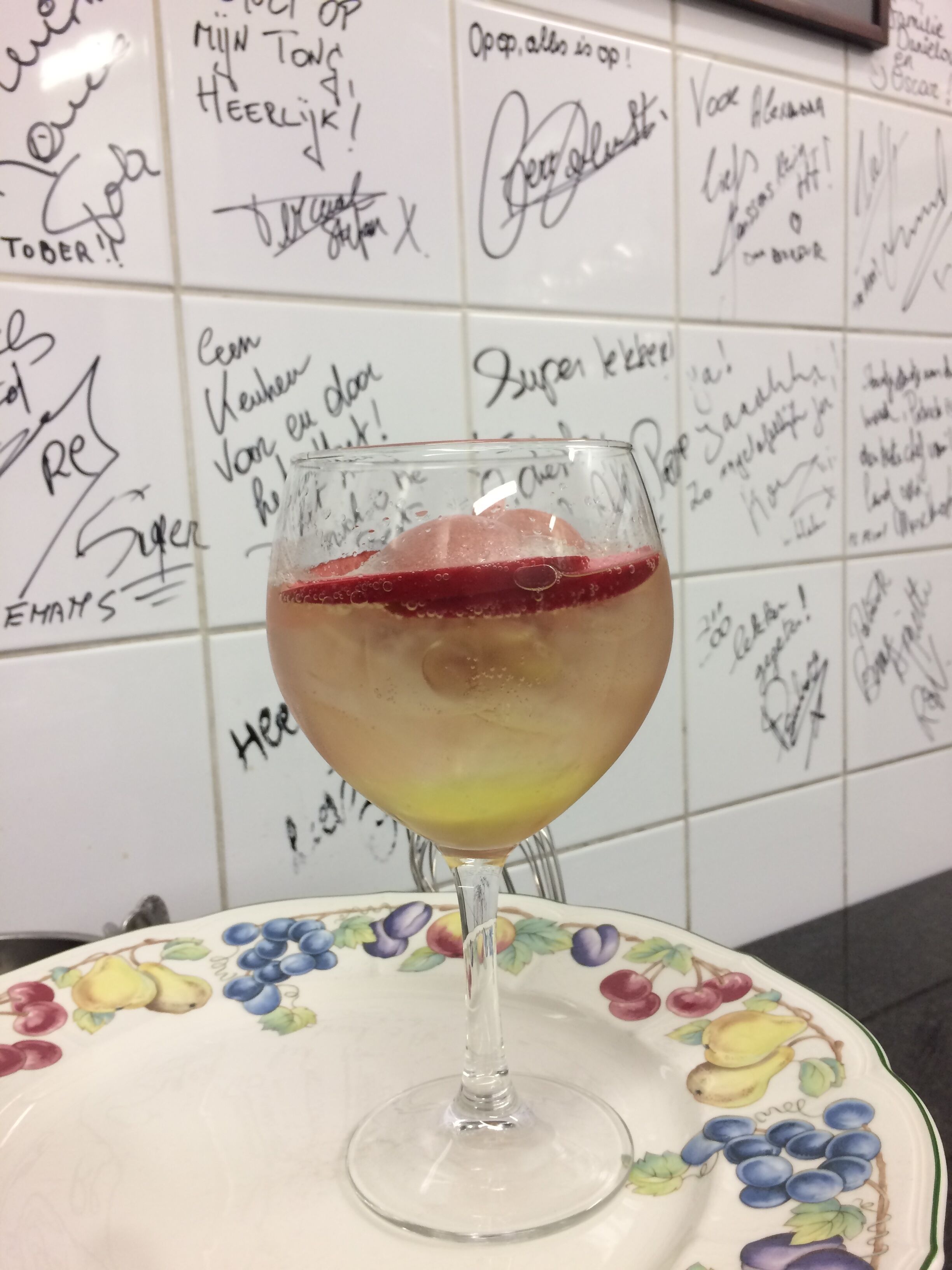 A glass of cocktail with a fruit garnish on a floral plate, set against a tiled wall with handwritten notes.