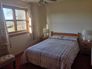 A bedroom with a bed, two bedside tables, a chair, a window with curtains, and a ceiling fan. - Balhousie Farm (Leven)