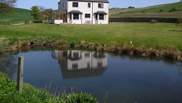 A two-story house with a large front porch, a pond in the foreground, and a grassy field in the background. - Balhousie Farm (Leven)