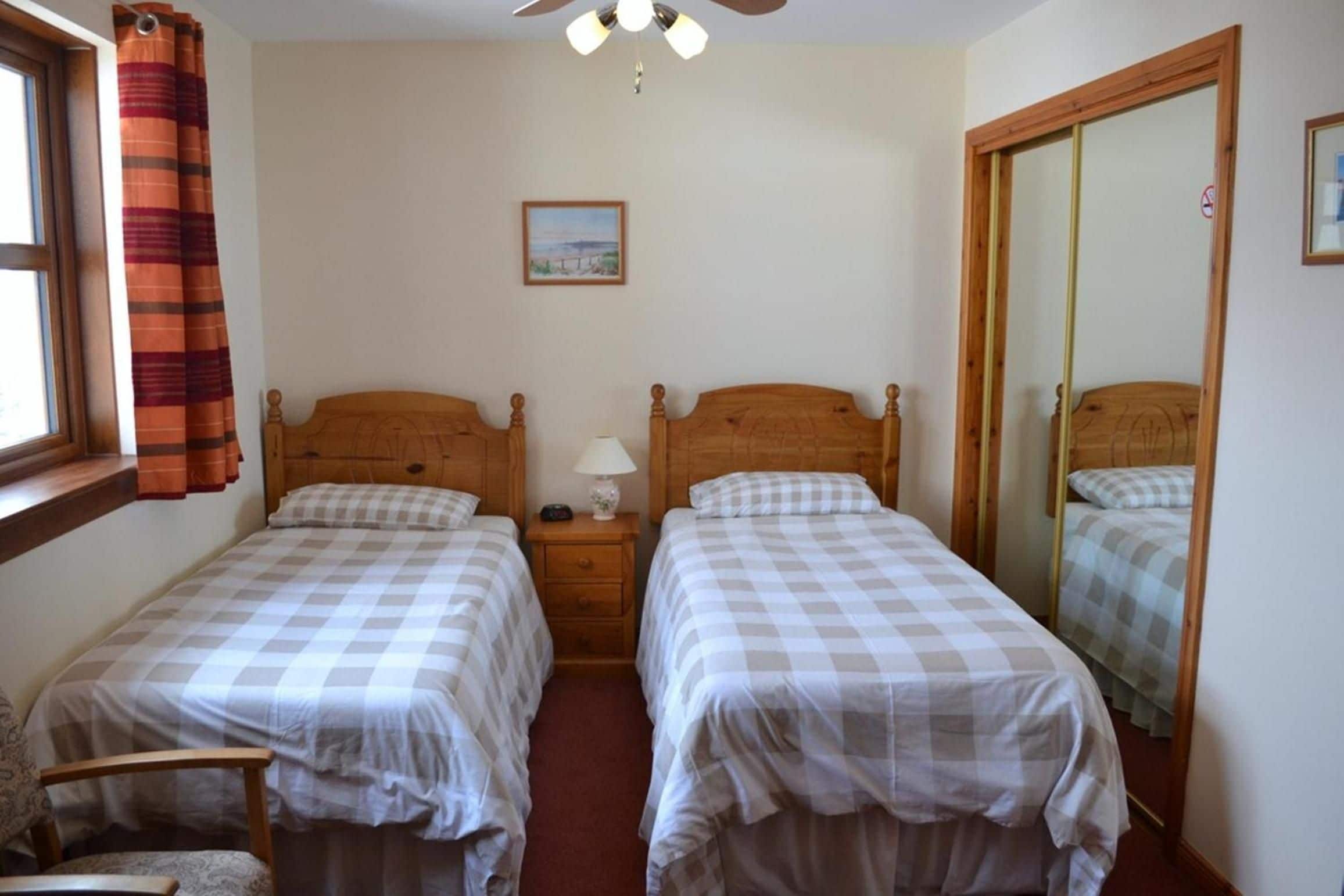 A bedroom with two single beds, a nightstand with a lamp, a window with curtains, a ceiling fan, and a framed picture on the wall.