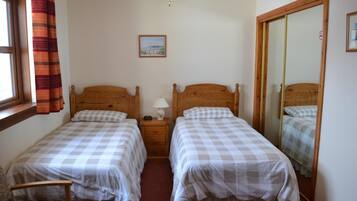 A bedroom with two single beds, a nightstand with a lamp, a window with curtains, a ceiling fan, and a framed picture on the wall.