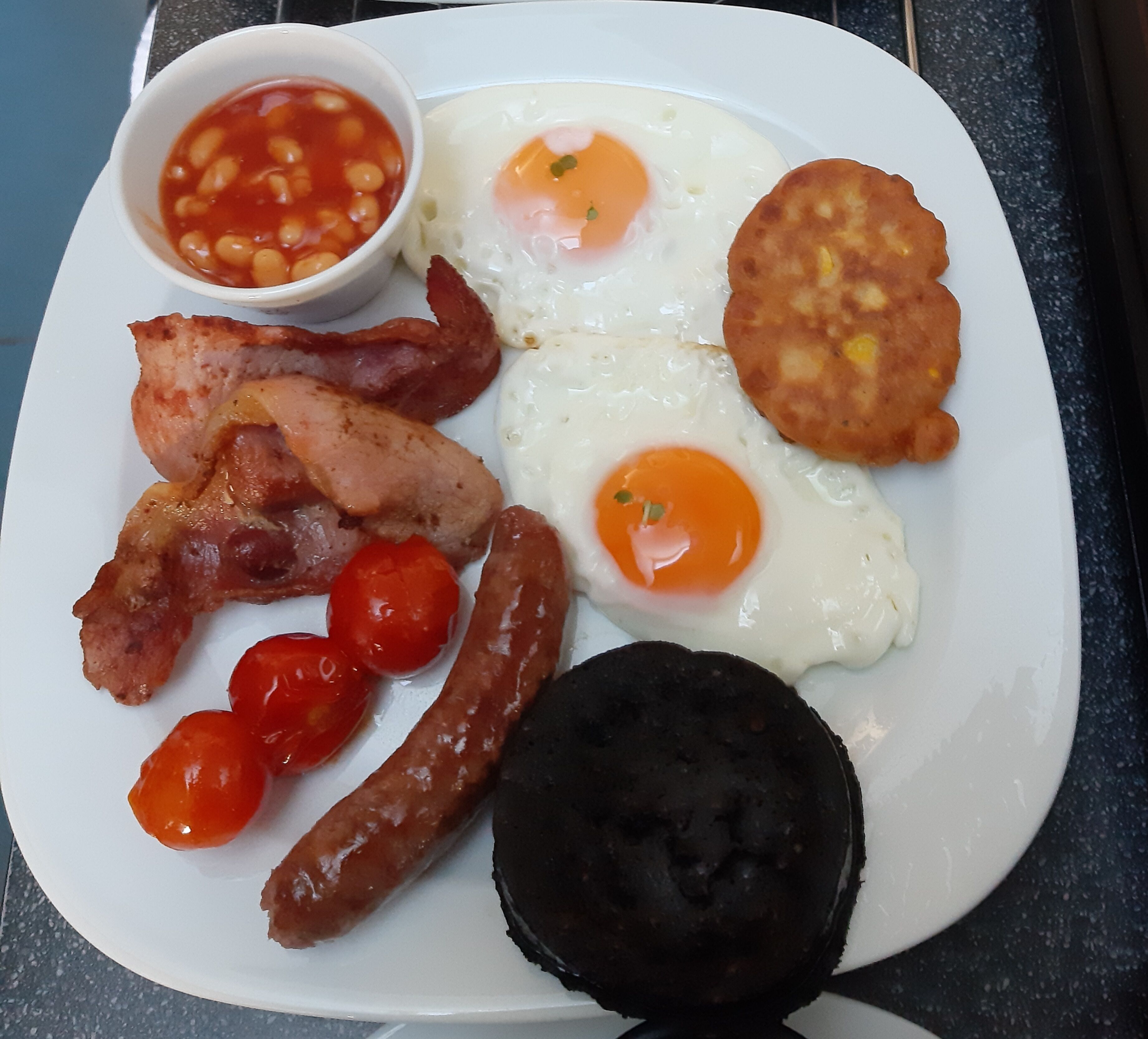 A plate of breakfast food including eggs, bacon, sausages, tomatoes, and a black pudding.