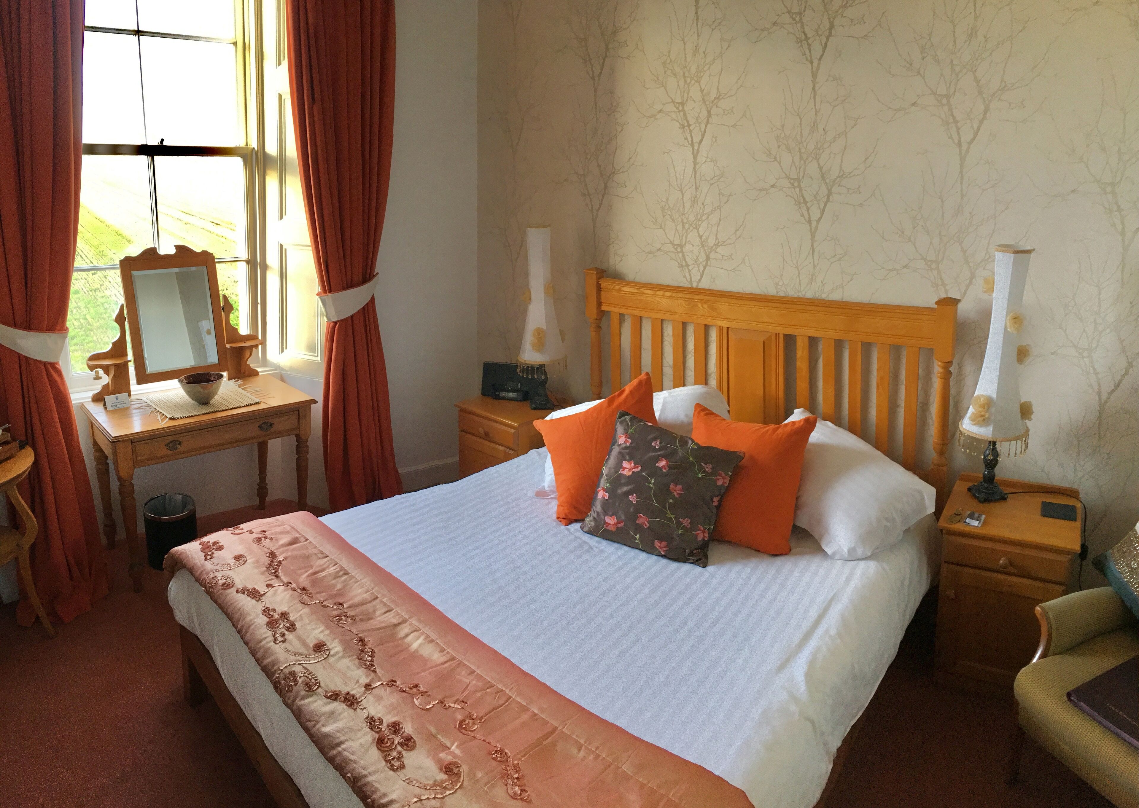 A neatly arranged bedroom with a bed, bedside tables, a nightstand, a lamp, a mirror, and a window with curtains.