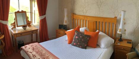A neatly arranged bedroom with a bed, bedside tables, a nightstand, a lamp, a mirror, and a window with curtains.