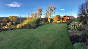 A well-maintained garden with a variety of plants and trees, a clear blue sky, and a red building in the background. - The Spindrift Guest House (Anstruther)