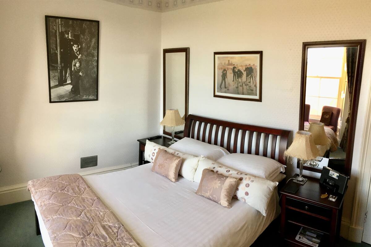 A neatly arranged hotel room with a large bed, two bedside tables, a nightstand, a mirror, and two framed artworks on the walls.