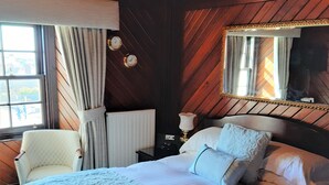 A bedroom with a large bed, a chair, a nightstand with a lamp, a mirror, and a window with curtains. - The Spindrift Guest House (Anstruther)