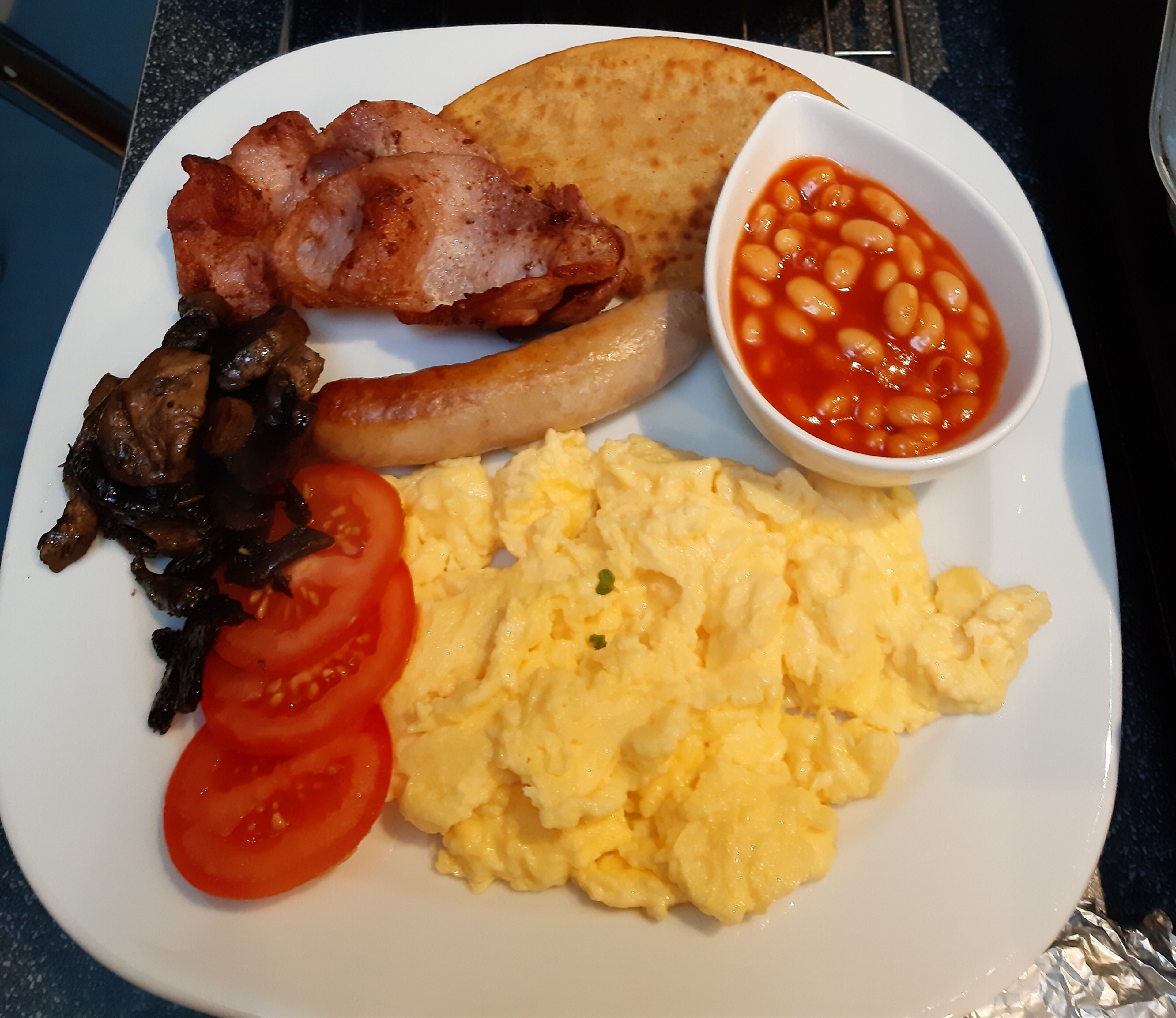 Un plato con huevos revueltos, tocino, salchichas, champiñones, tomates, tostadas y frijoles horneados.