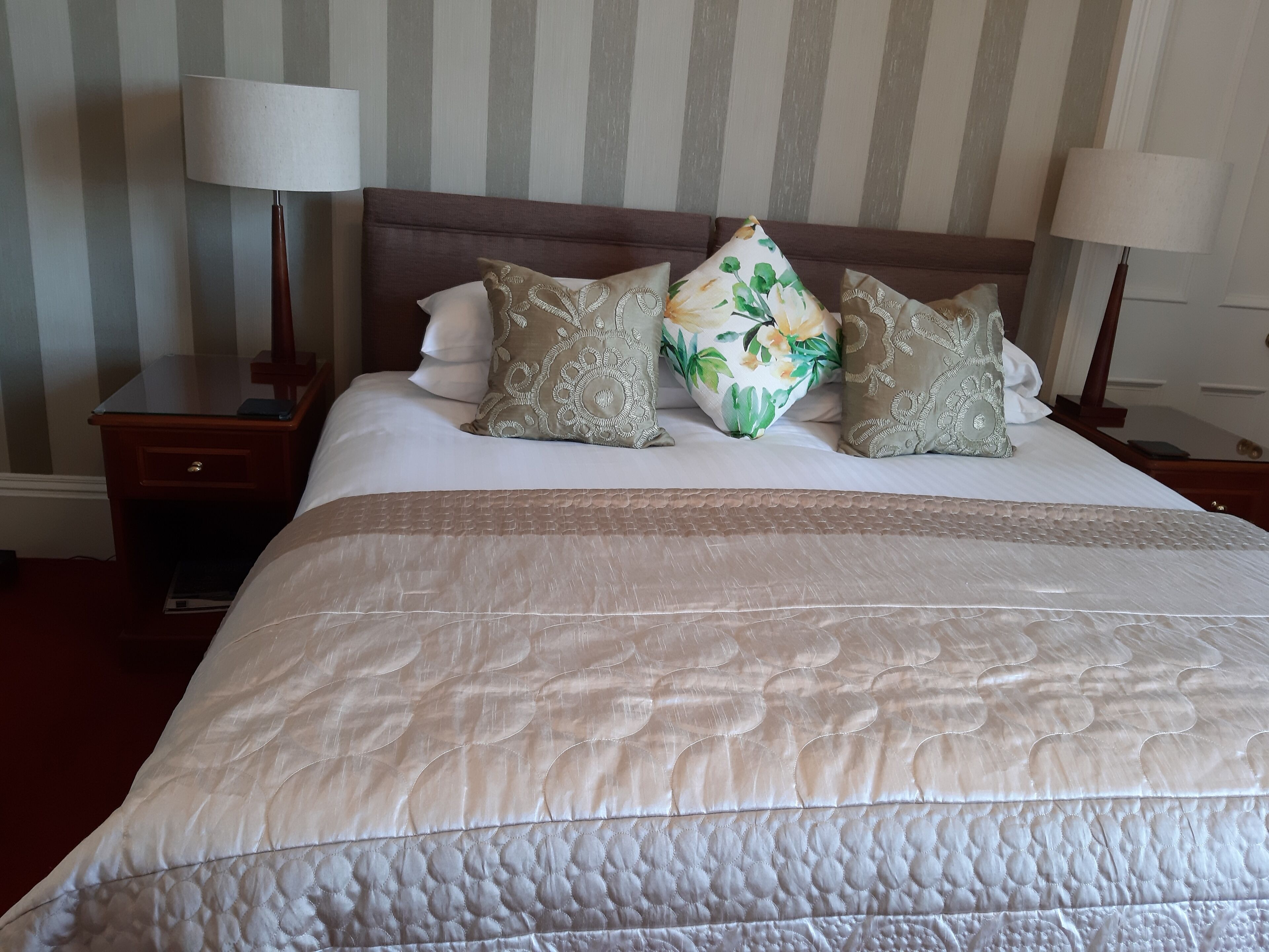 A neatly made bed with a quilted headboard, two bedside tables with lamps, and a striped wall in the background.