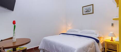 A hotel room with a bed, a bedside table with a lamp, a chair with a floral cushion, a small table with a vase of flowers, and a TV mounted on the wall.