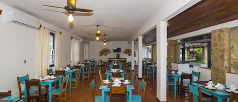 Uma sala de jantar espaçosa com várias mesas e cadeiras, ventiladores de teto e vista para o exterior.