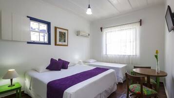 Habitación de hotel con dos camas, una mesita, una silla, un ventanal con cortinas, un televisor montado en la pared y un marco de fotos.