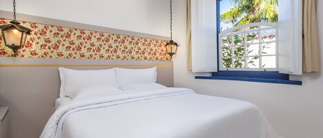 Una cama bien hecha en una habitación con papel tapiz floral, un colgador de techo y una ventana con vista a palmeras.