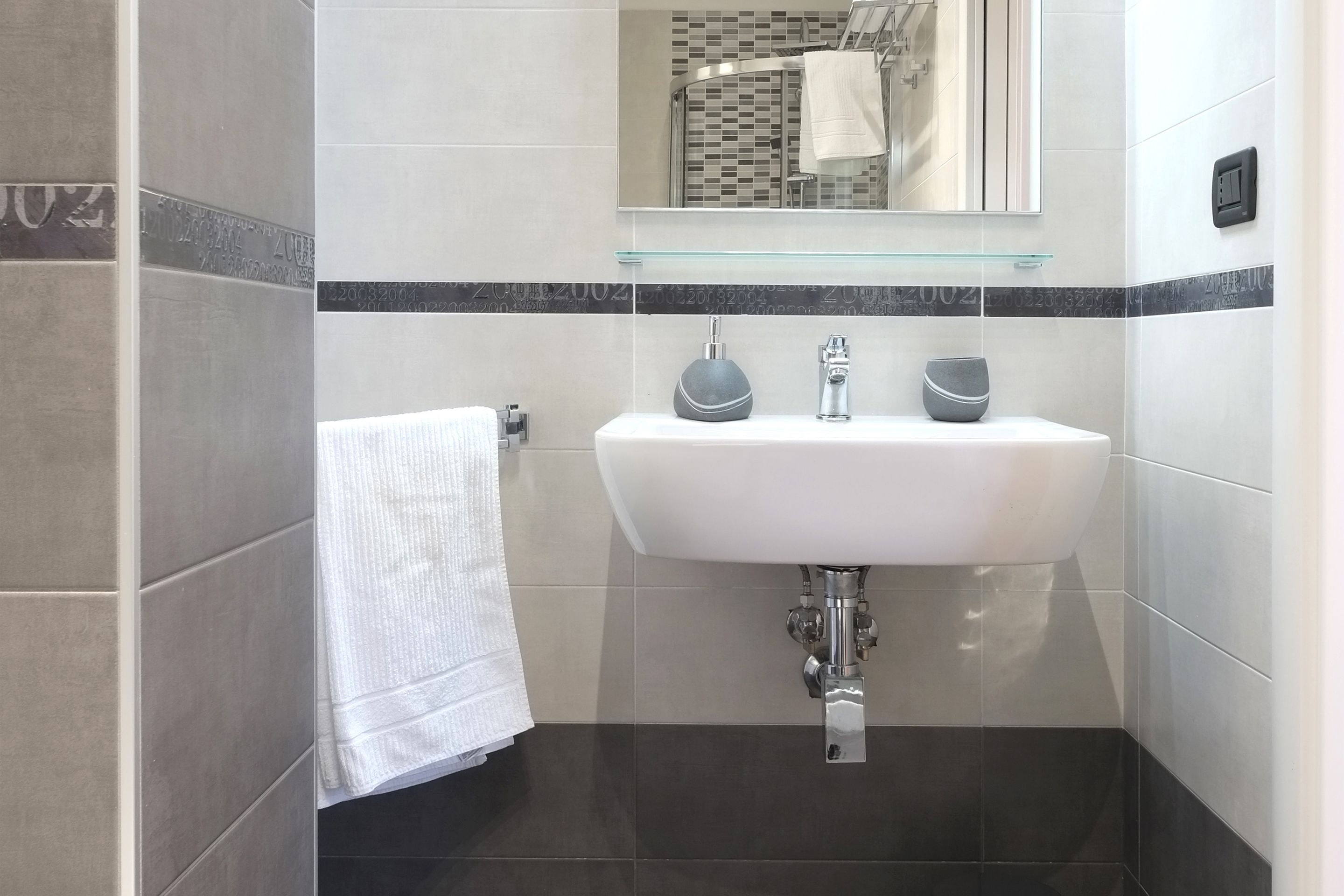Un baño moderno con un lavamanos blanco, dos dispensadores de jabón, un estante de vidrio sobre el lavamanos y una toalla colgada en la pared.