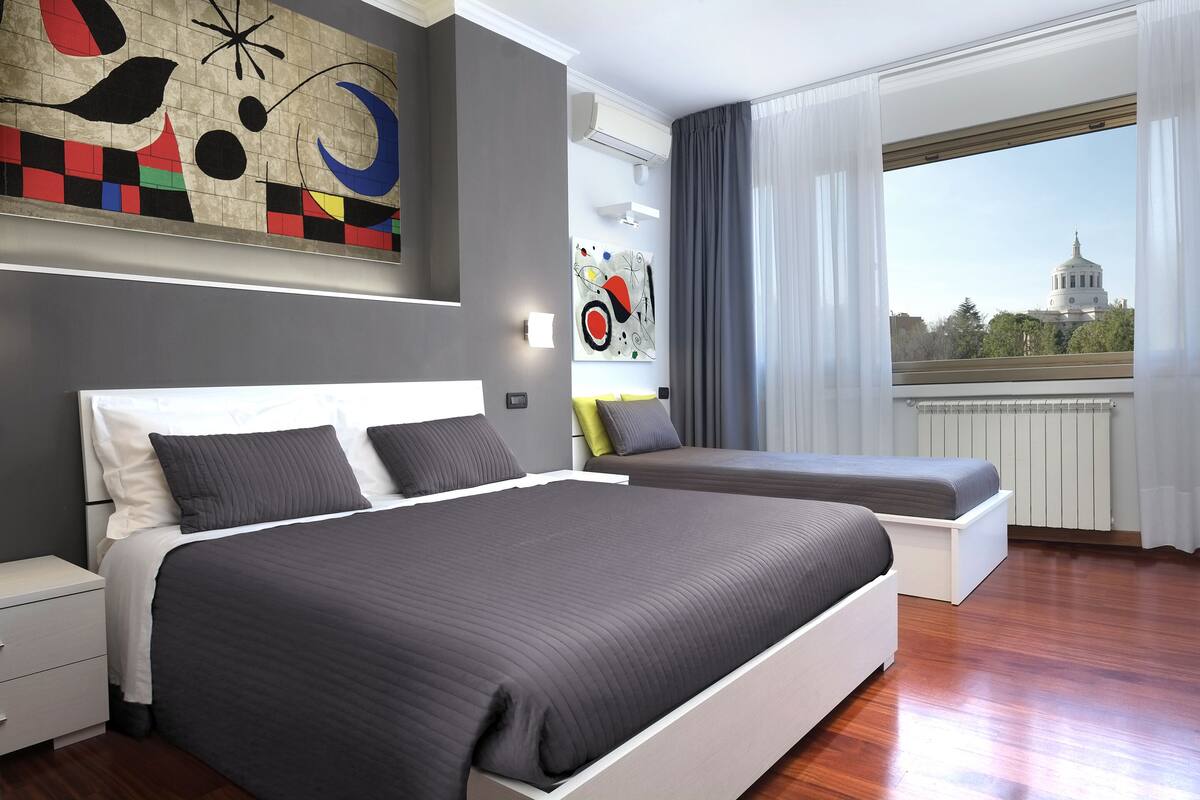 Una habitación de hotel moderna con una cama grande, una cama más pequeña, una mesita de noche y vistas a un edificio con cúpula a través de la ventana.