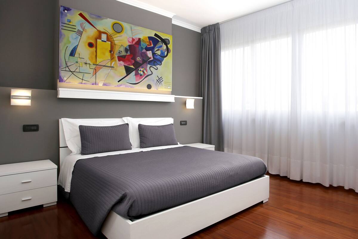 Un dormitorio moderno con una cama grande, una mesita de noche, un cuadro en la pared y una ventana con cortinas.