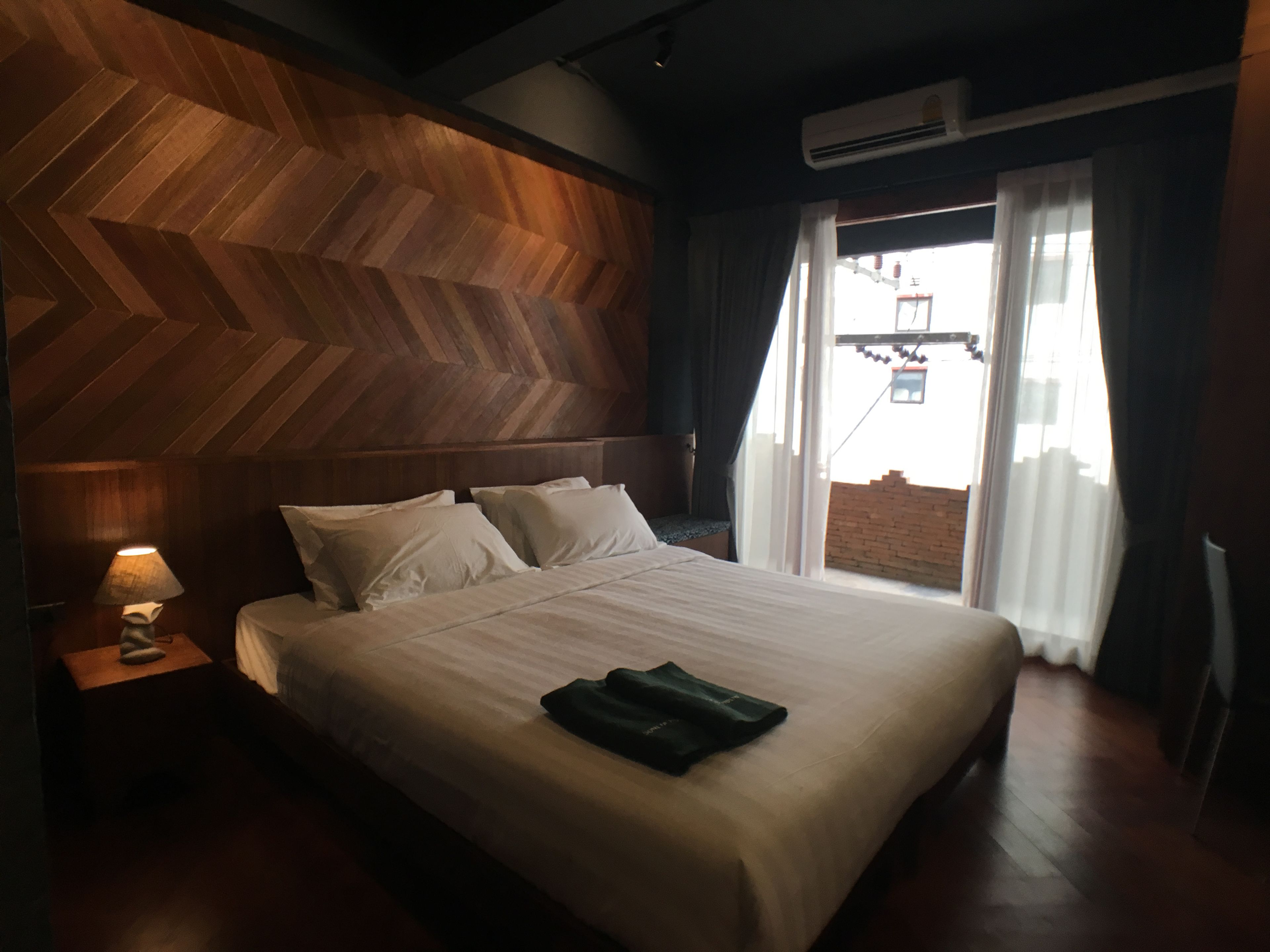 Un cuarto de hotel con una cama grande, paneles de madera en las paredes, una mesita de noche con lámpara y vista a un muro de ladrillos.