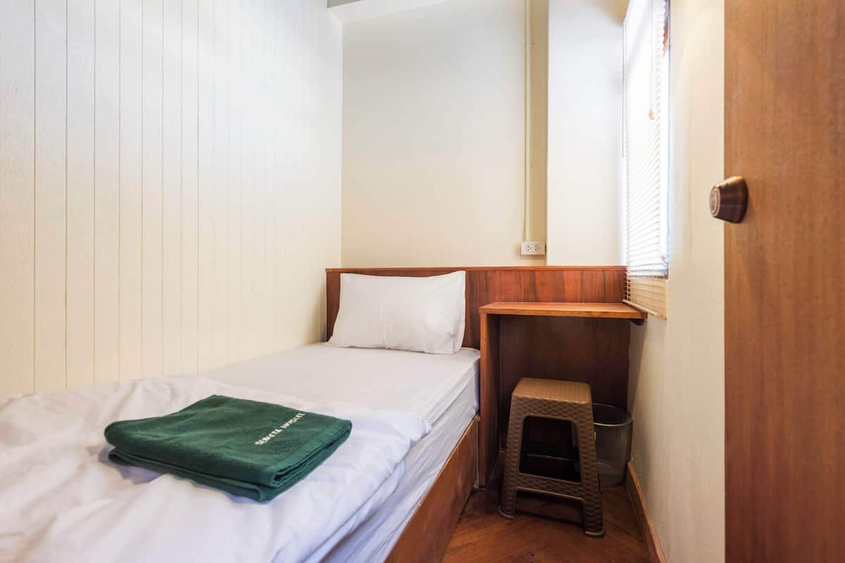 Een kleine kamer met een enkel bed, een houten hoofdbord, een klein krukje en een raam met jaloezieën.