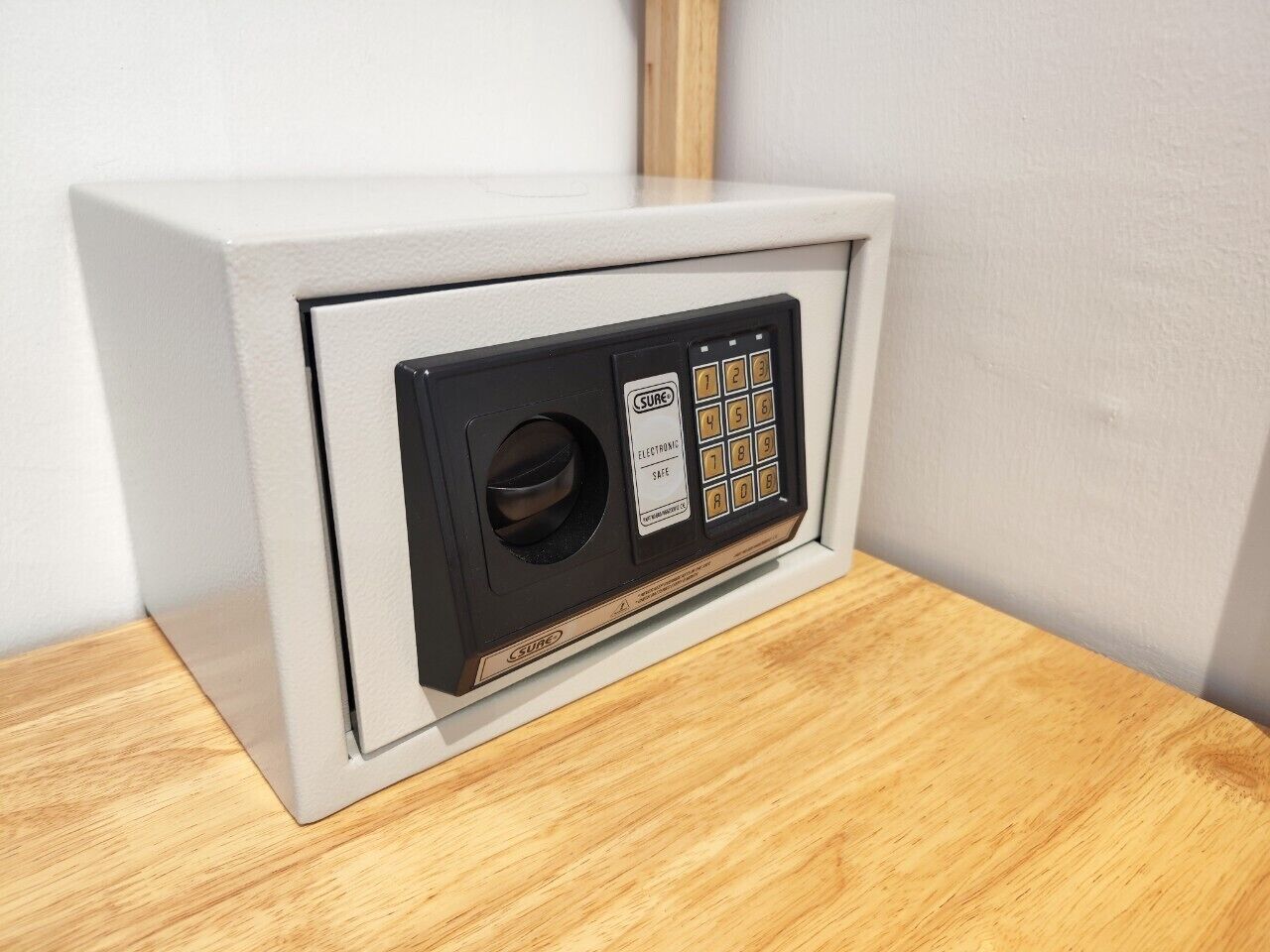 A safe with a keypad and a slot for a key, placed in a corner against a white wall.