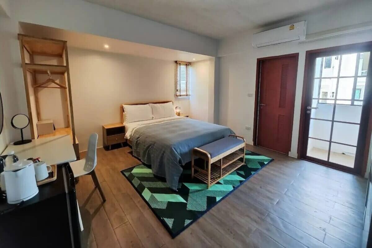 Een slaapkamer met een bed, nachtkastje, stoel en een bureau met stoel. Er is een raam met jaloezieën, een deur en uitzicht op buiten.