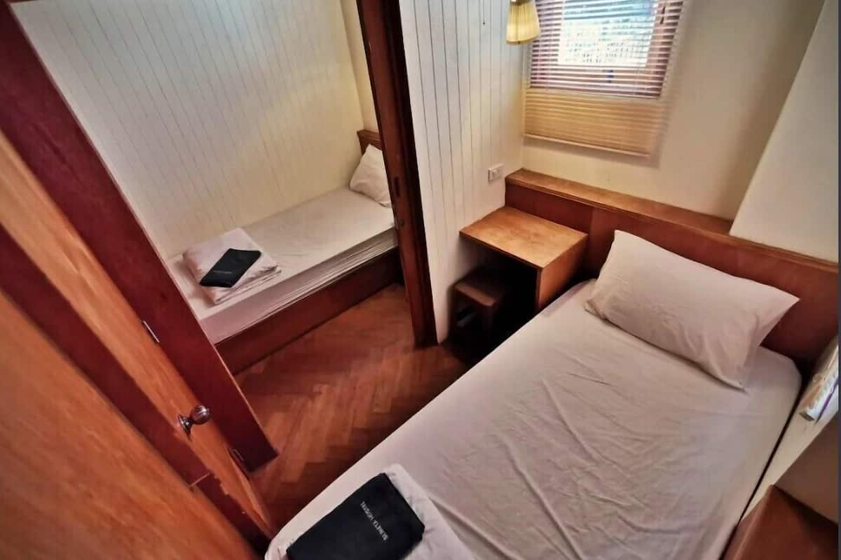 Een kleine slaapkamer met een enkel bed, een klein nachtkastje, een lamp en een raam met jaloezieën.