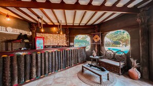 A rustic bar area with a wooden counter, a Coca-Cola refrigerator, and a view of a pool through a window.