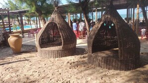 Two woven, conical structures on a sandy beach, likely used for seating or storage, with a pool and umbrellas in the background.