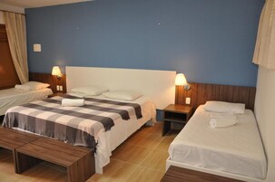 Habitación de hotel con dos camas individuales, una cama grande, cabecero de madera, mesitas de noche con lámparas y un banco.