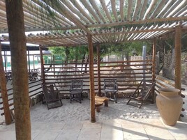 A covered outdoor seating area with wooden tables and chairs, a large woven basket, and a large ceramic pot.