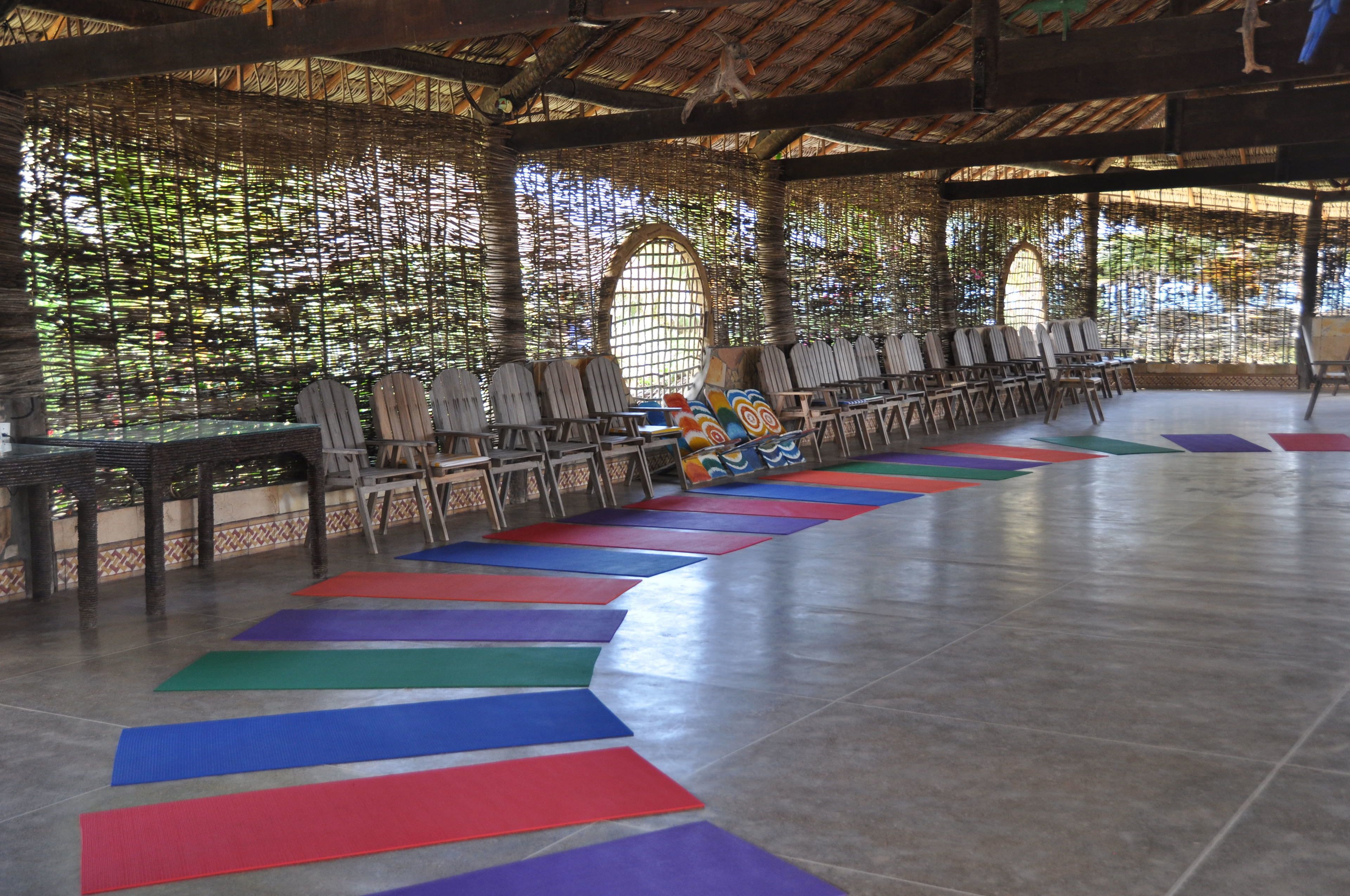 A covered outdoor area with a tiled floor, rows of chairs, and colorful mats arranged in a line.