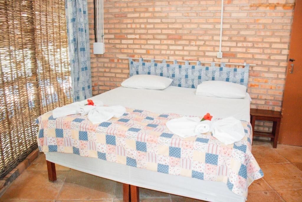 Uma cama bem arrumada com colcha e toalhas, uma parede de tijolos e uma pequena mesa de cabeceira.