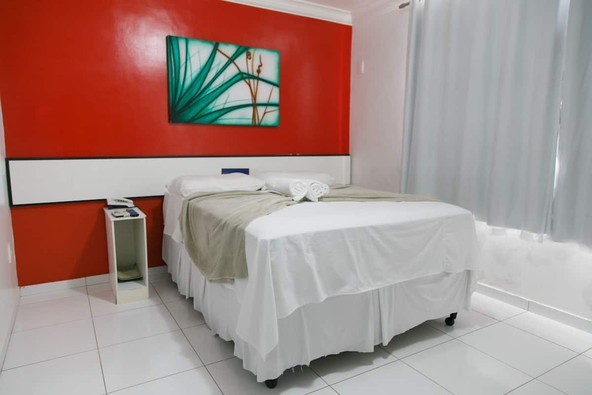Una habitació d'hotel amb un llit, una tauleta de nit, un quadre a la paret i una finestra amb cortines.