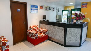 Une salle d’attente avec une porte en bois, un canapé rouge à motifs floraux, un comptoir avec un ordinateur et un téléphone, des certificats encadrés accrochés au mur, et un panneau indiquant que le Wi-Fi est gratuit.