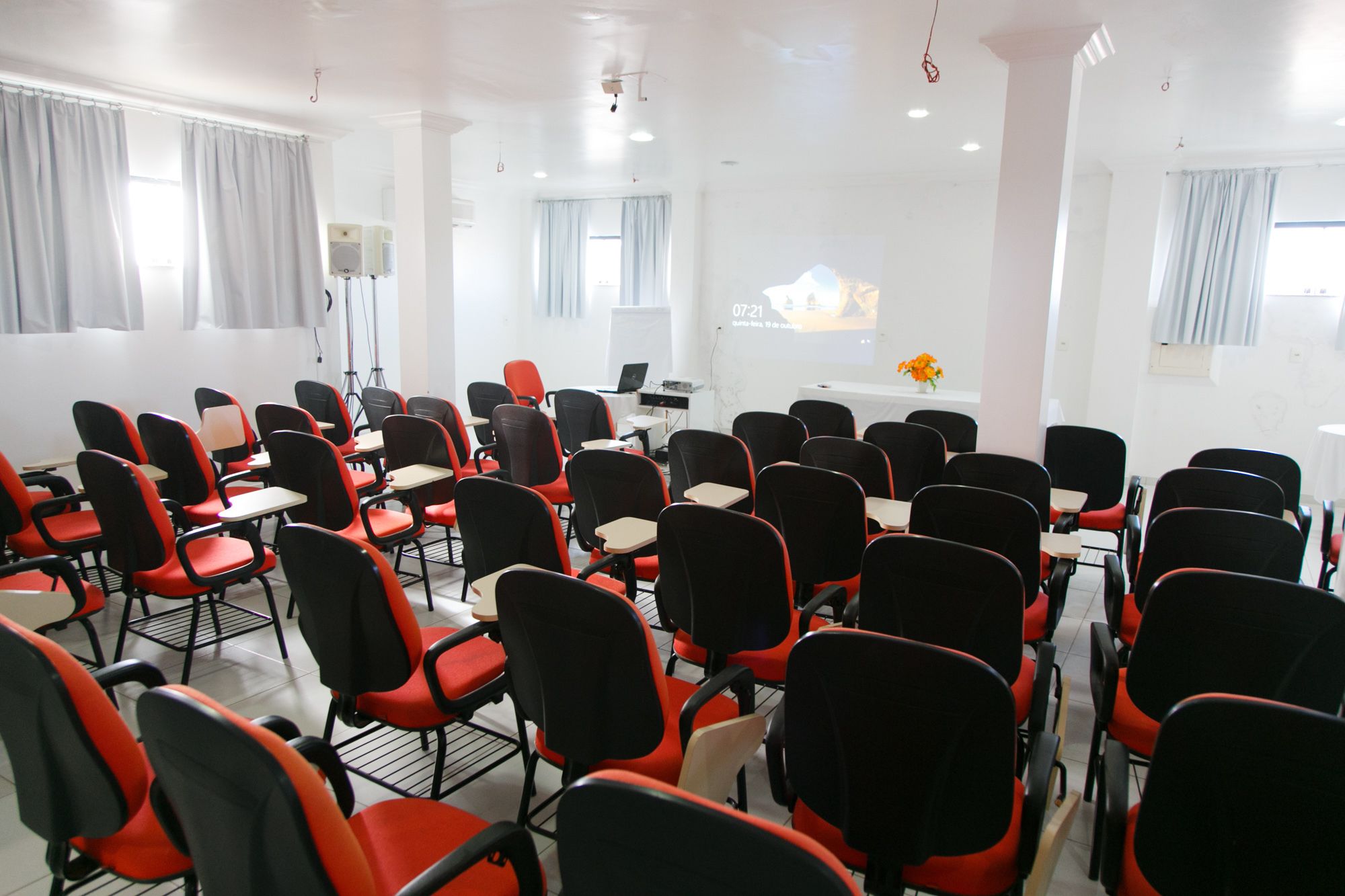 Ein Konferenzraum mit Reihen schwarzer und roter Stühle, einer Projektionsleinwand und einem Laptop auf einem Tisch.