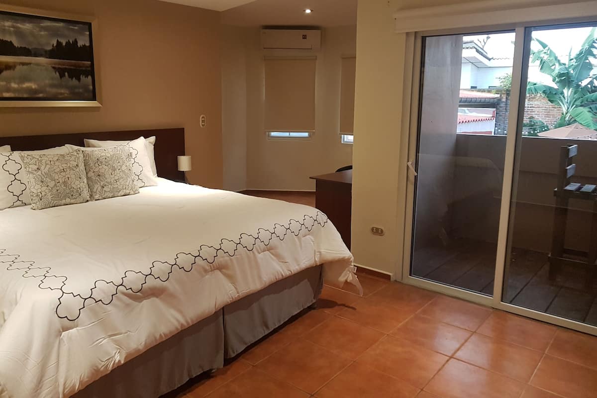 A hotel room with a large bed, a desk, a chair, and a sliding glass door leading to an outdoor area.