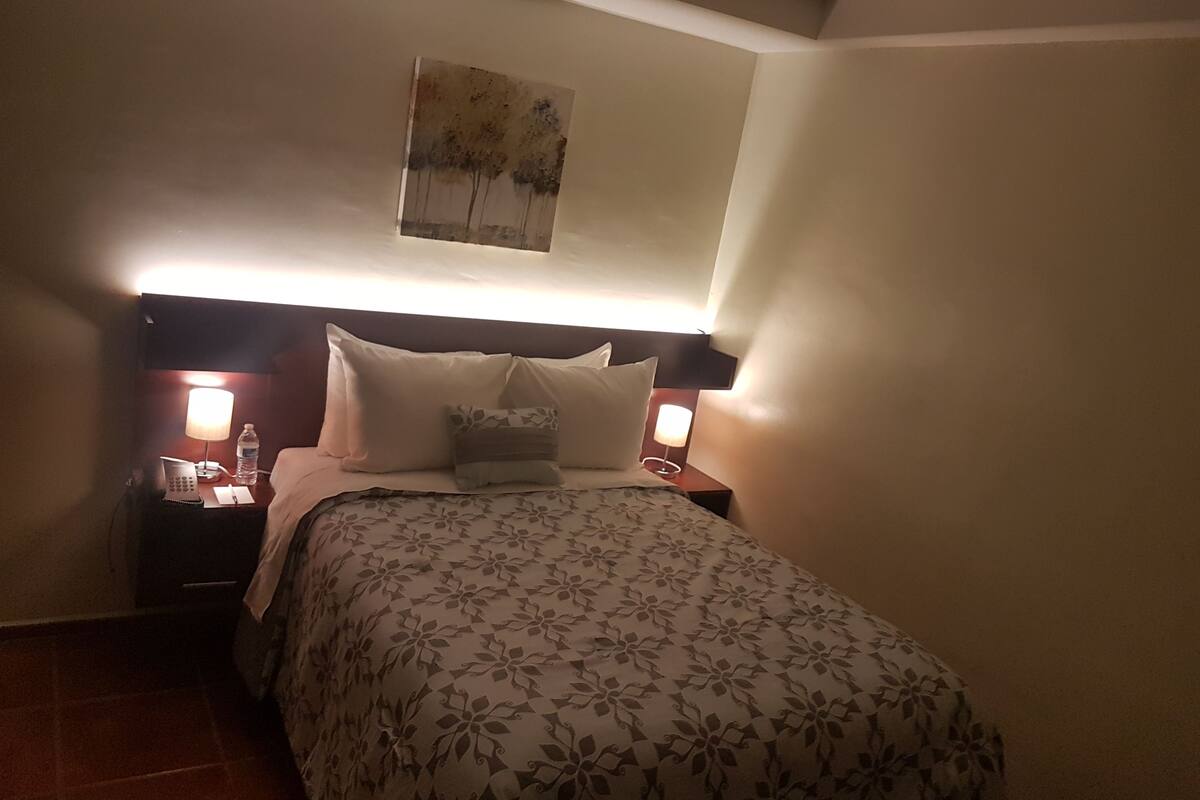 A hotel room with a bed, two bedside lamps, a nightstand, a painting on the wall, and a tiled floor.