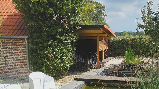 A garden with a pond, a wooden structure, and a brick building. - Gite L'archipel (Ellezelles)