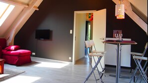 A loft-style room with a sloped ceiling, a red sofa, a TV mounted on the wall, and a dining area with chairs and a table. - Gite L'archipel (Ellezelles)