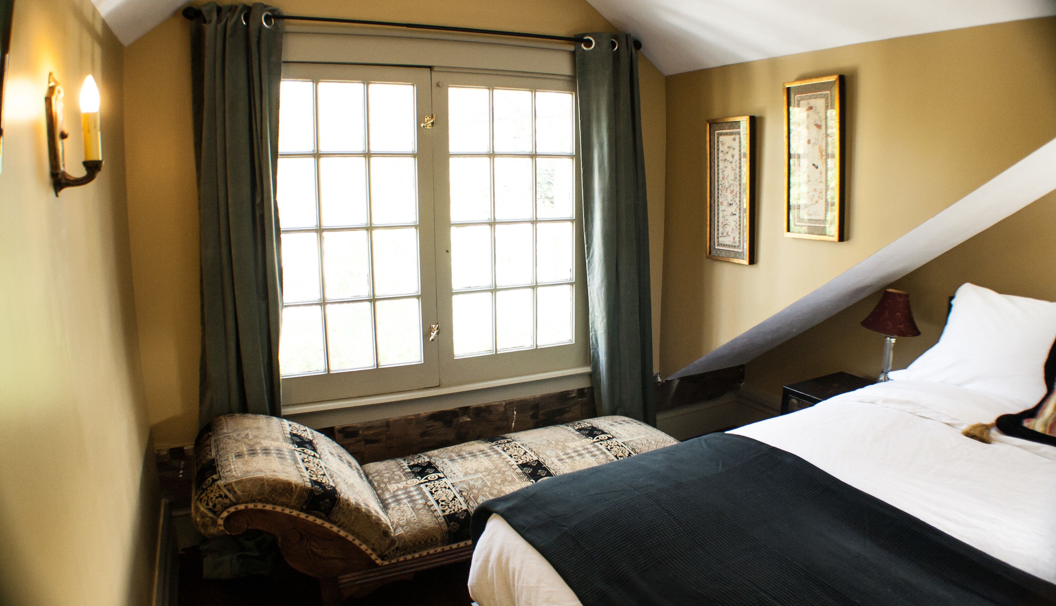 A bedroom with a bed, a window with curtains, a bedside table with a lamp, and two framed pictures on the wall.