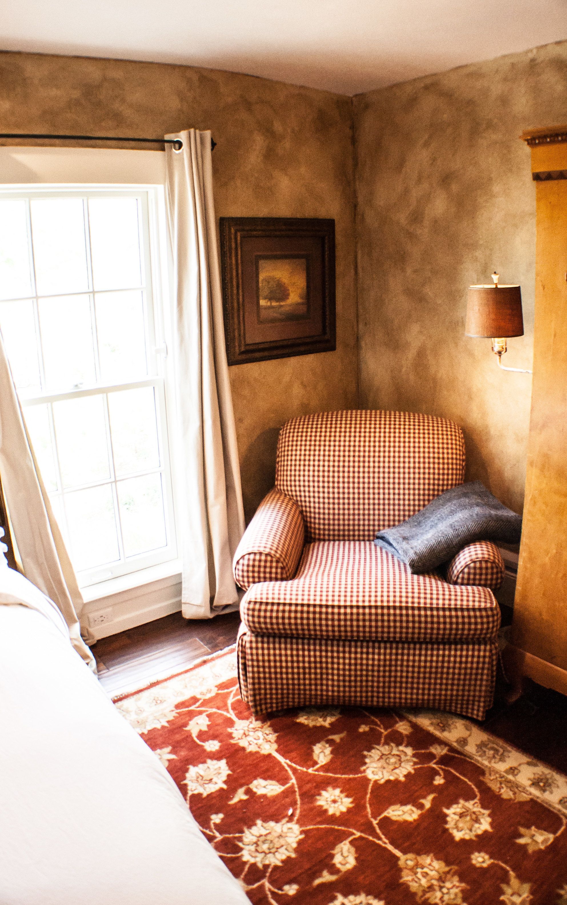 Une pièce avec un fauteuil à motifs à carreaux, un tapis à motifs, une fenêtre avec des rideaux et une lampe fixée au mur.
