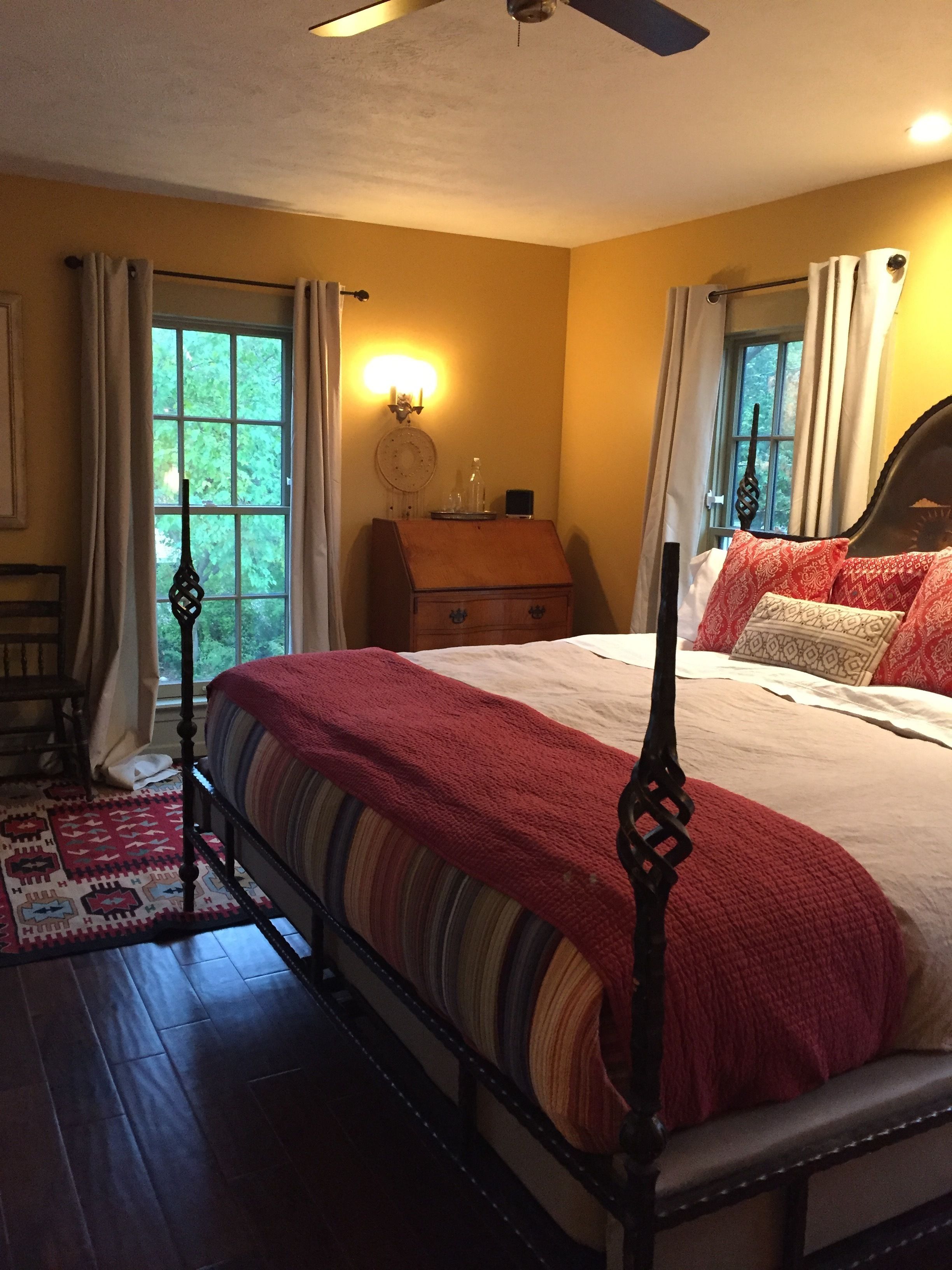 A bedroom with a four-poster bed, a wooden nightstand, a wall-mounted lamp, and a view of greenery through the window.